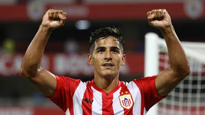 A Puebla player in a red and white striped jersey, celebrating with arms raised after scoring a goal in the Estadio...