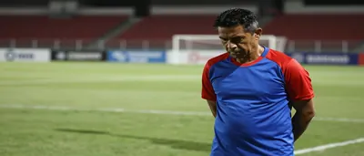 A Puebla coach, mid-40s, wearing a plain blue and red training top with clean unbroken fabric across the chest, stands...