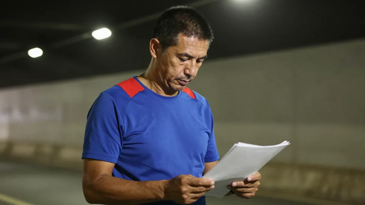 A 35-year-old male coach with a rugged build, standing in a dimly lit tunnel, his face illuminated by the faint glow of a...