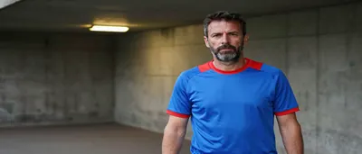 A focused coach, mid-30s, with a weathered face and a wispy beard, stands in the tunnel leading to the pitch, his blue...