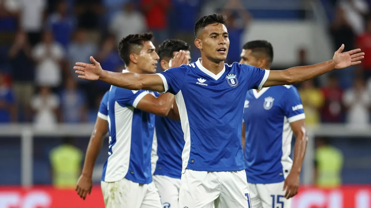 A 25-year-old Puebla player, wearing a blue and white jersey, celebrates a goal with his teammates, with a stadium crowd...