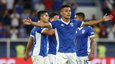 A 25-year-old Puebla player, wearing a blue and white jersey, celebrates a goal with his teammates, with a stadium crowd...