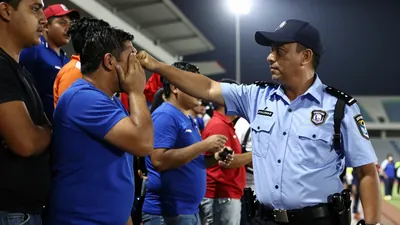 A security guard in a blue uniform, with a white shirt and black pants, raises his fist to punch a Puebla fan in the...