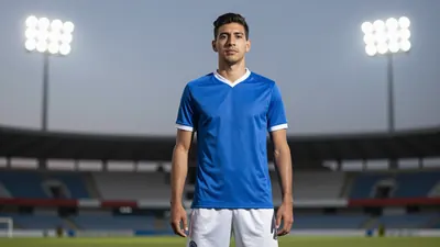 A photorealistic image of an anonymous Puebla player in a blue jersey with white shorts, standing in front of a stadium...