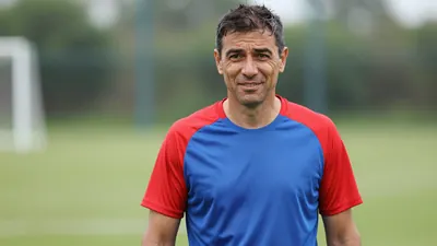 Middle-aged male coach, mid-40s, athletic build, waist-up, wearing a plain blue and red training top (solid blue front...
