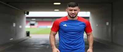 A muscular young man in a plain blue and red training top, mid-30s, with a tense expression, standing in a floodlit...