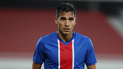 Mid-30s Puebla midfielder, lean, dark hair, intense eyes, shoulders hunched, wearing a plain blue jersey with red...